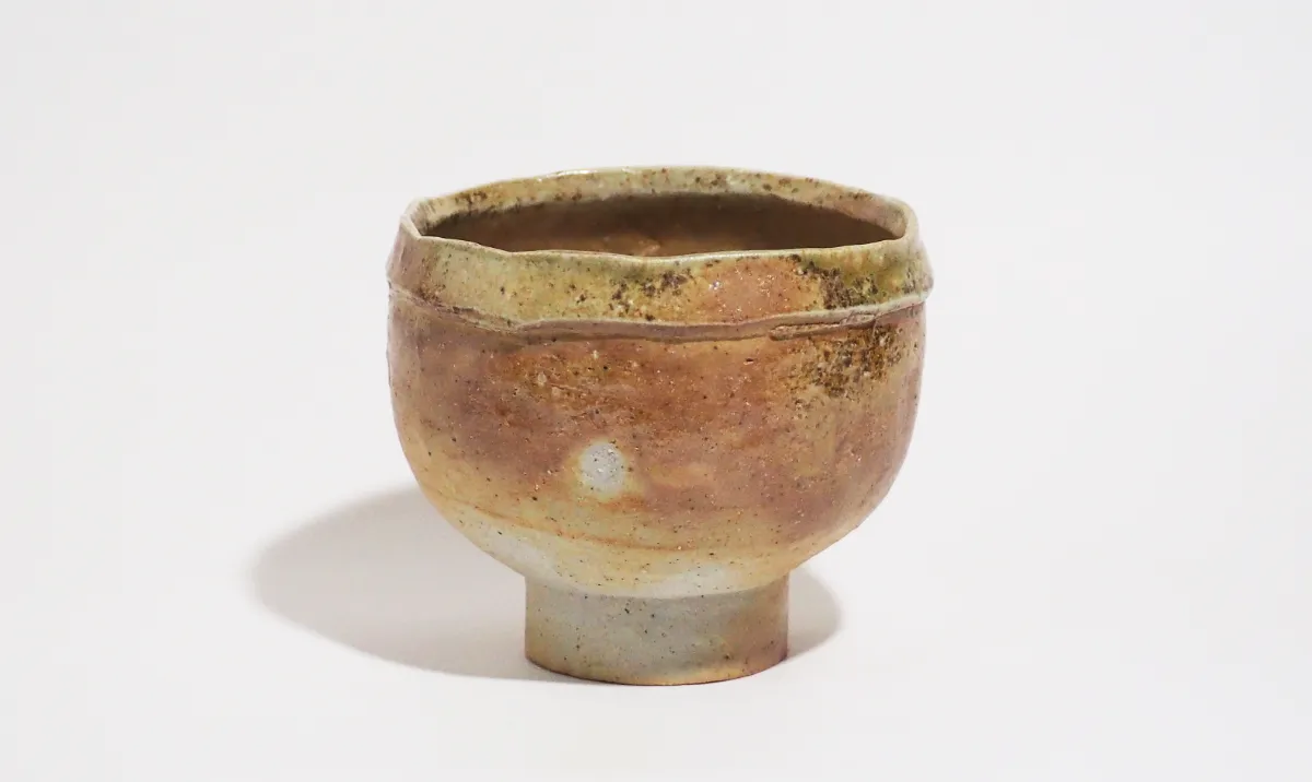 黄褐色を基調とした抹茶茶碗で、表面には自然なまだら模様が現れている。側面は柔らかく丸みを帯びた形状をしており、全体に落ち着いた立ち姿の印象を与える。下部の高台は灰白色で、本体との色の対比が特徴的。口縁付近には折り返し状の造形が施され、手捻りならではの立体感と緊張感を加えている。整った輪郭と、偶然性のある表情が組み合わされた作品。