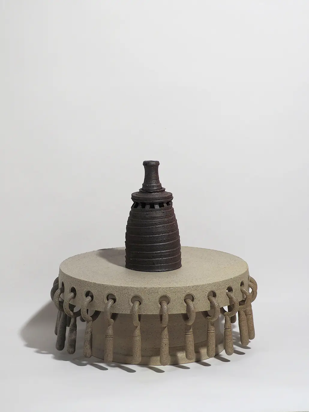 二つの陶芸作品で構成されたセット作品。上部作品は黒褐色の塔状作品で、全体は上に向かって緩やかに細くなる形をしている。側面には螺旋状の溝が巡り、上部には覗き窓を思わせる小さな開口状の造形が施されている。最上部はごく短い円筒形となっており、塔全体に静かな緊張感を与えている。下部作品は灰白色の台座作品で、円盤状の天板を円筒形の支柱が支える構造になっている。天板の側面には多数の穴が開けられており、それぞれに短い鎖やイヤリングのような装飾が吊り下げられている。装飾はわずかに揺れ動く構造となっており、静的な塔作品との対比を生み出している。全体として、重厚な塔と動きのある台座が組み合わされた立体作品となっている。